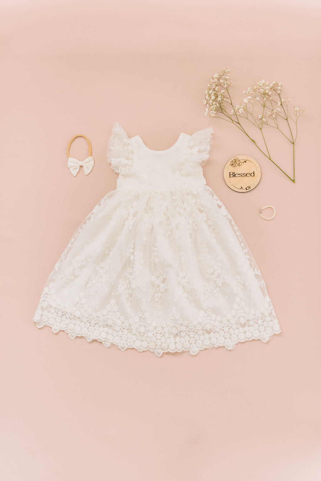 Flat lay of lace dress with flutter sleeves, white bow, wooden blessed sign and pearl bracelet.