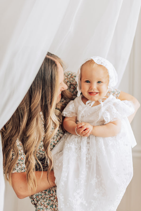Mom making baby girl smile in lace white dress with lace bonnet.