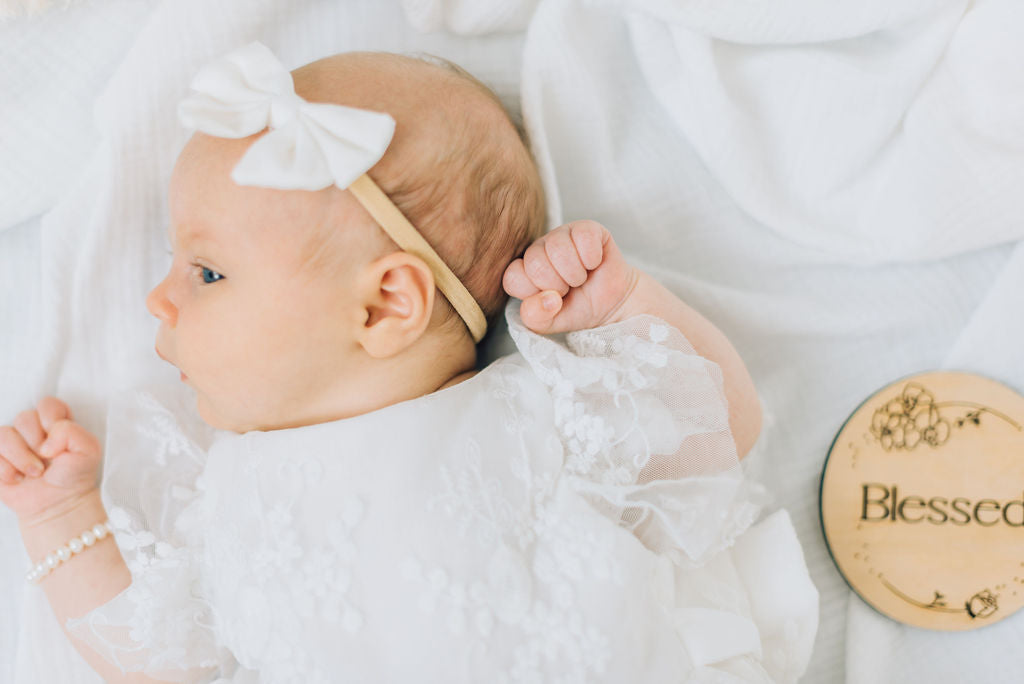 Close up view of baby in lace dress with ruffle sleeves, blessed wooden sign, white bow and pearl bracelet.