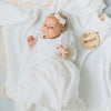 Baby girl lahing on white blanket with cream tassels in her white lace blessing dress. 