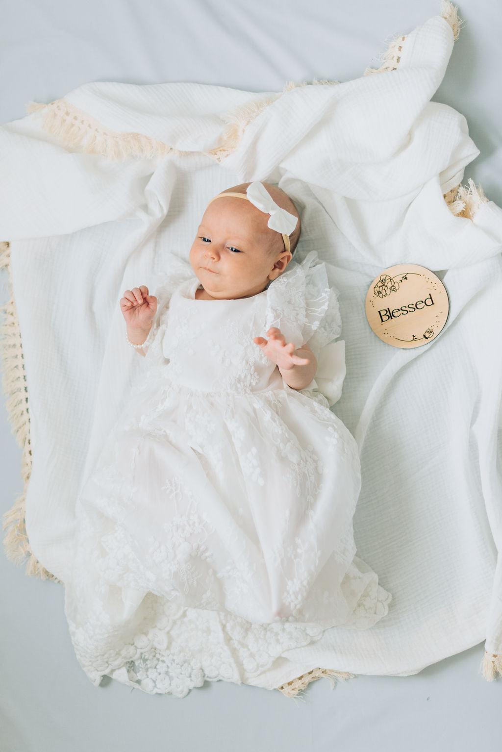 Baby girl lahing on white blanket with cream tassels in her white lace blessing dress. 