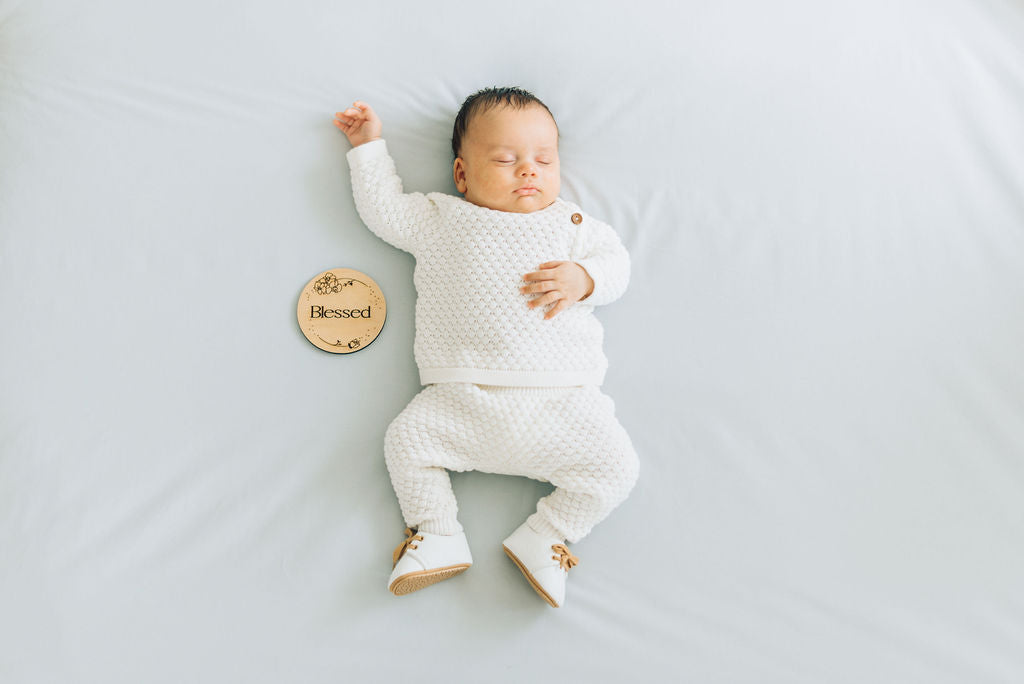 Baby sleeping in white set next to Blessed wooden sign. 