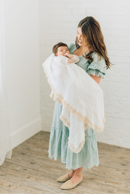 White blanket with cream tassels on edge wrapped around baby boy in mothers arms.