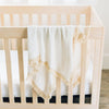 White baby blanket with tassels draped over wooden crib.