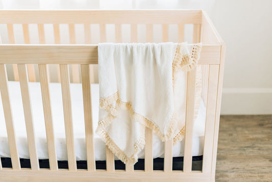 White baby blanket with tassels draped over wooden crib.