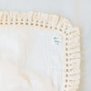 Detail shot of white blanket with cream tassels with Miriia tag next to greenery. 