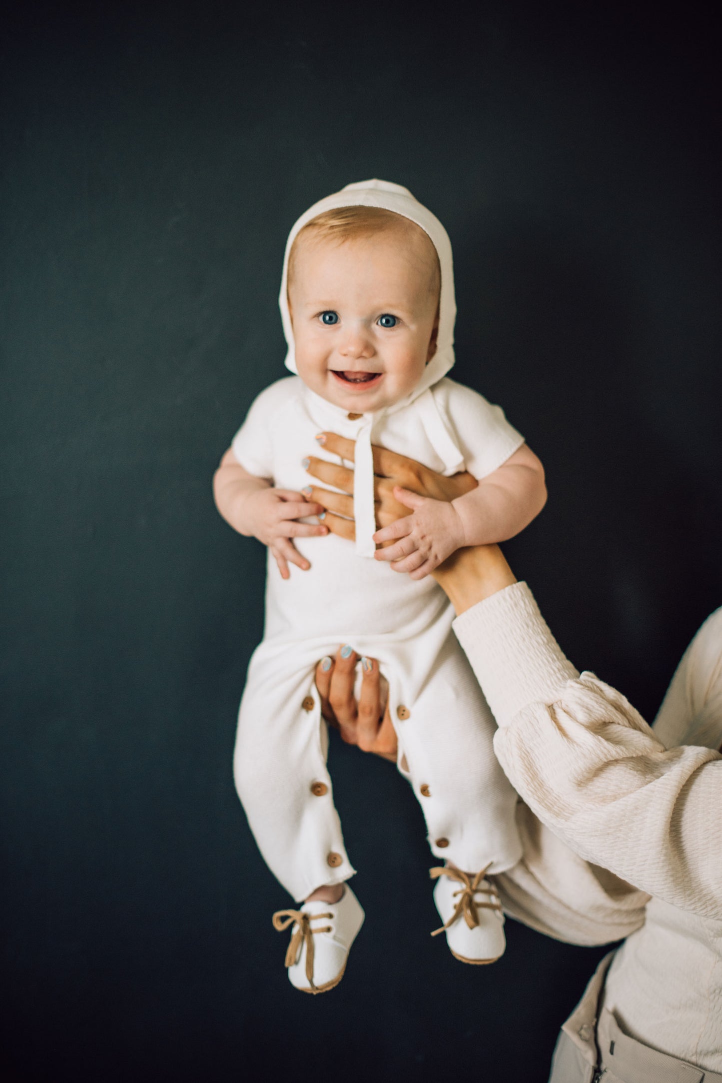 Baby boy being help up in white onesie, white bonnet, and white shoes with brown laces.