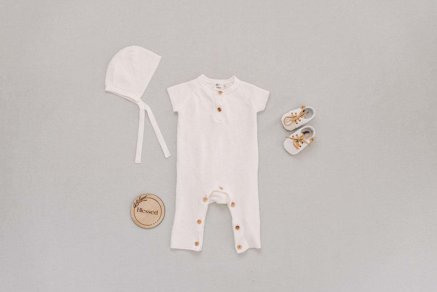Flat lat with white bonnet, while onesie, white shoes with brown laces and a wooden blessed sign.