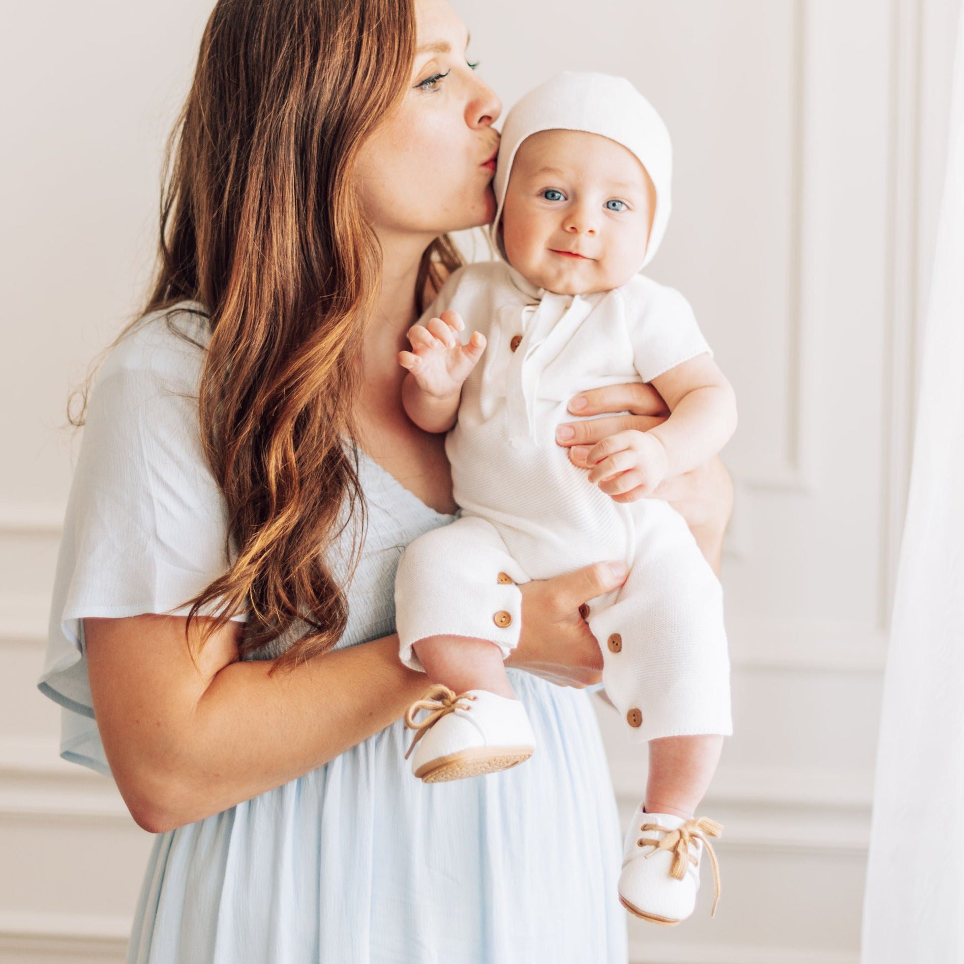 Mom lovingly kissing baby boy in white one piece sweater outfit