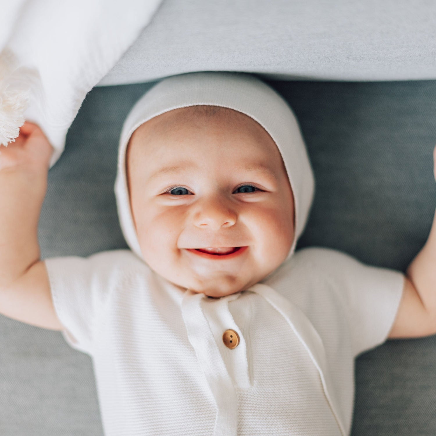 Smiling baby boy in white blessing outfit with white bonnet next to white blanket with tassels.