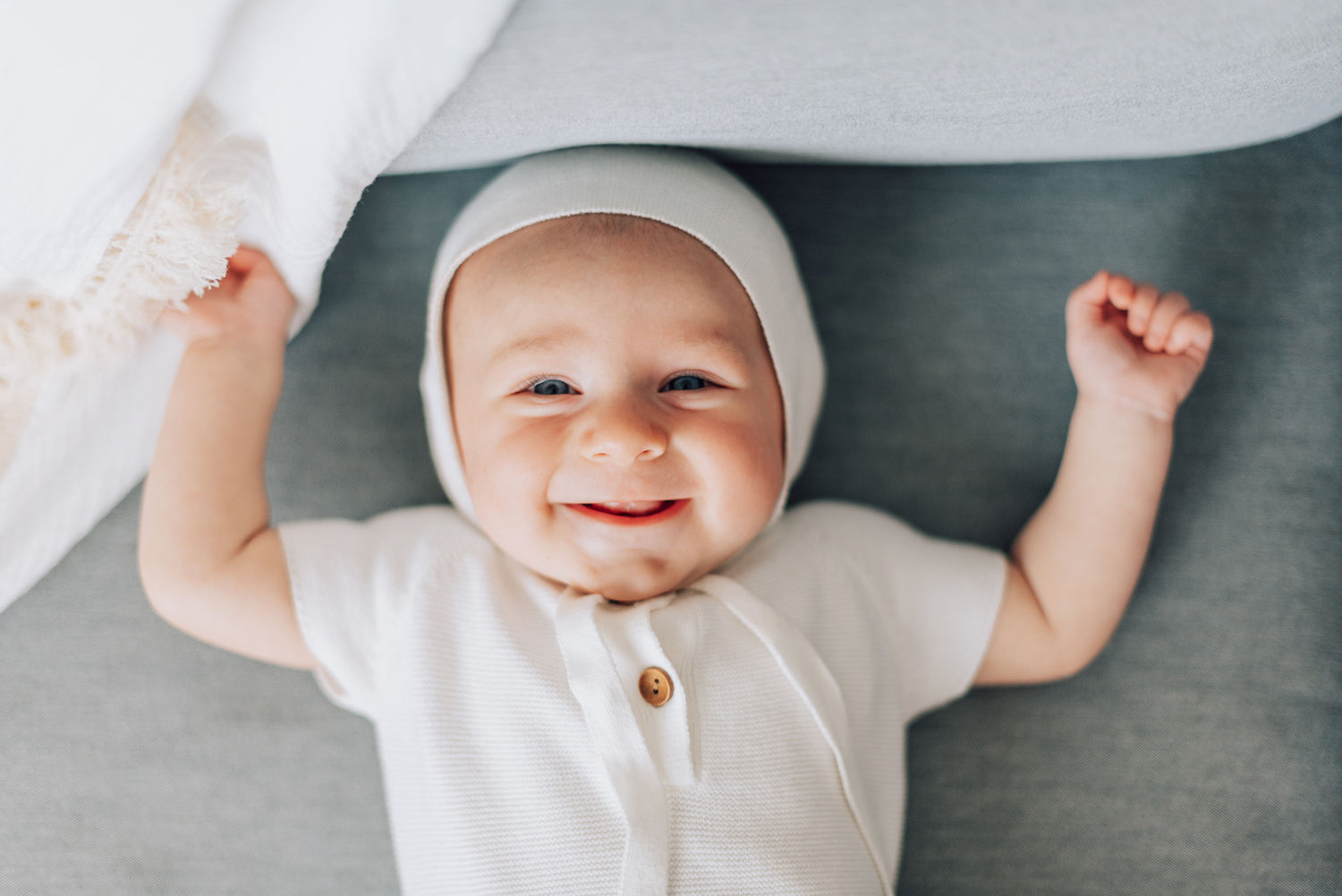 Smiling baby boy in white blessing outfit with white bonnet next to white blanket with tassels.
