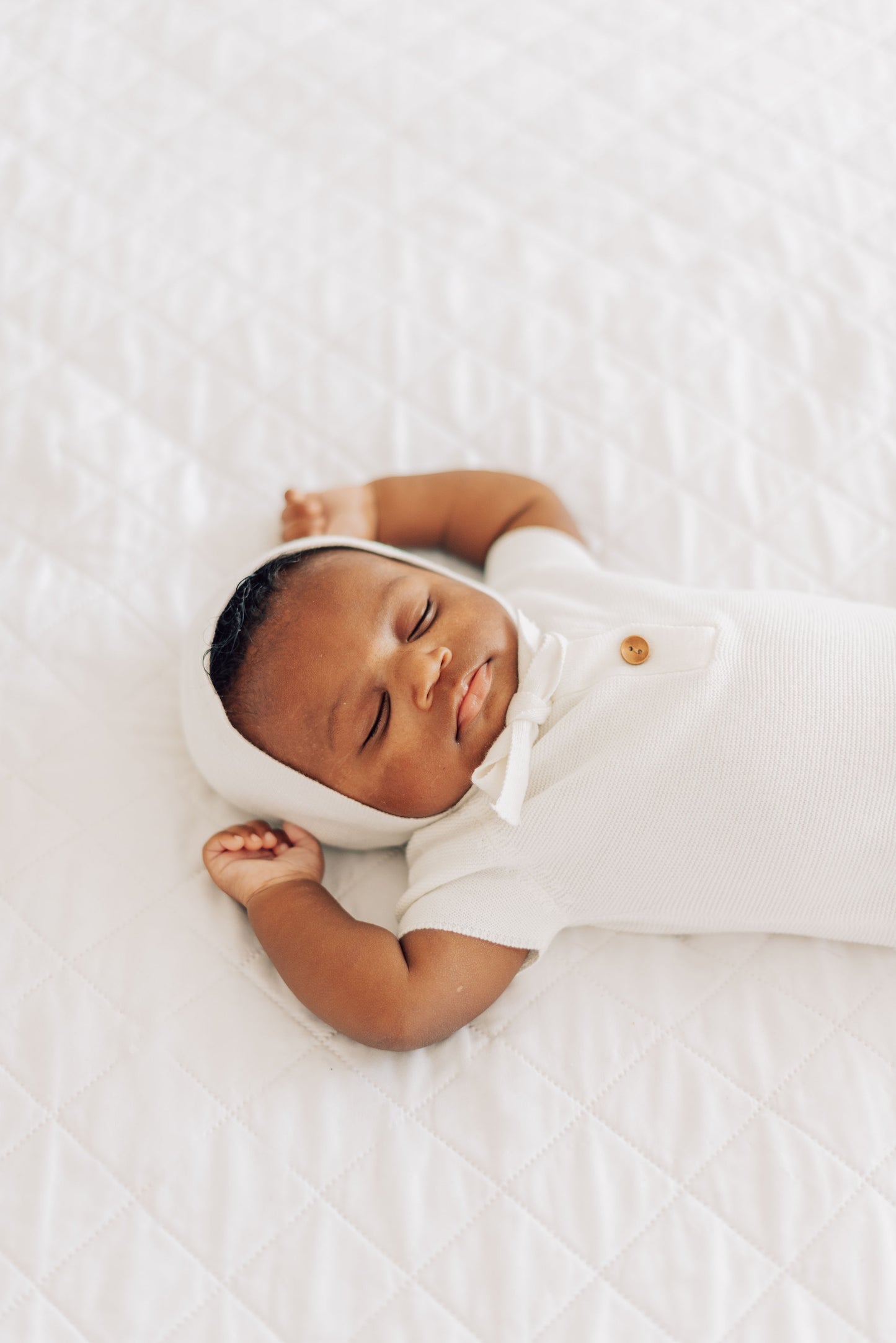 Baby laying on white comforter in white bonnet and white outfit with brown buttons.