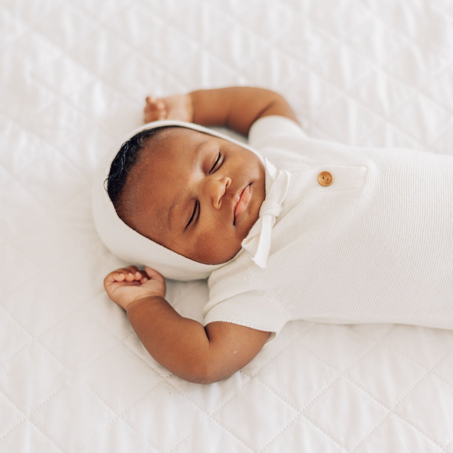 Baby laying on white comforter in white bonnet and white outfit with brown buttons.