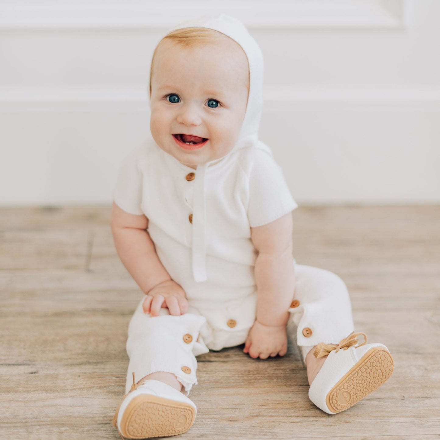 Baby boy sitting up on the ground smiling in a white blessing outfit.